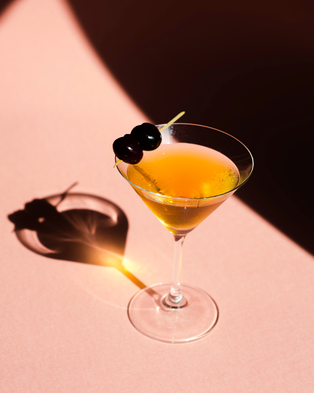 long-stem cocktail glass filled with a brown liquid and garnished with 2 brandied cherries sat on a pink surface