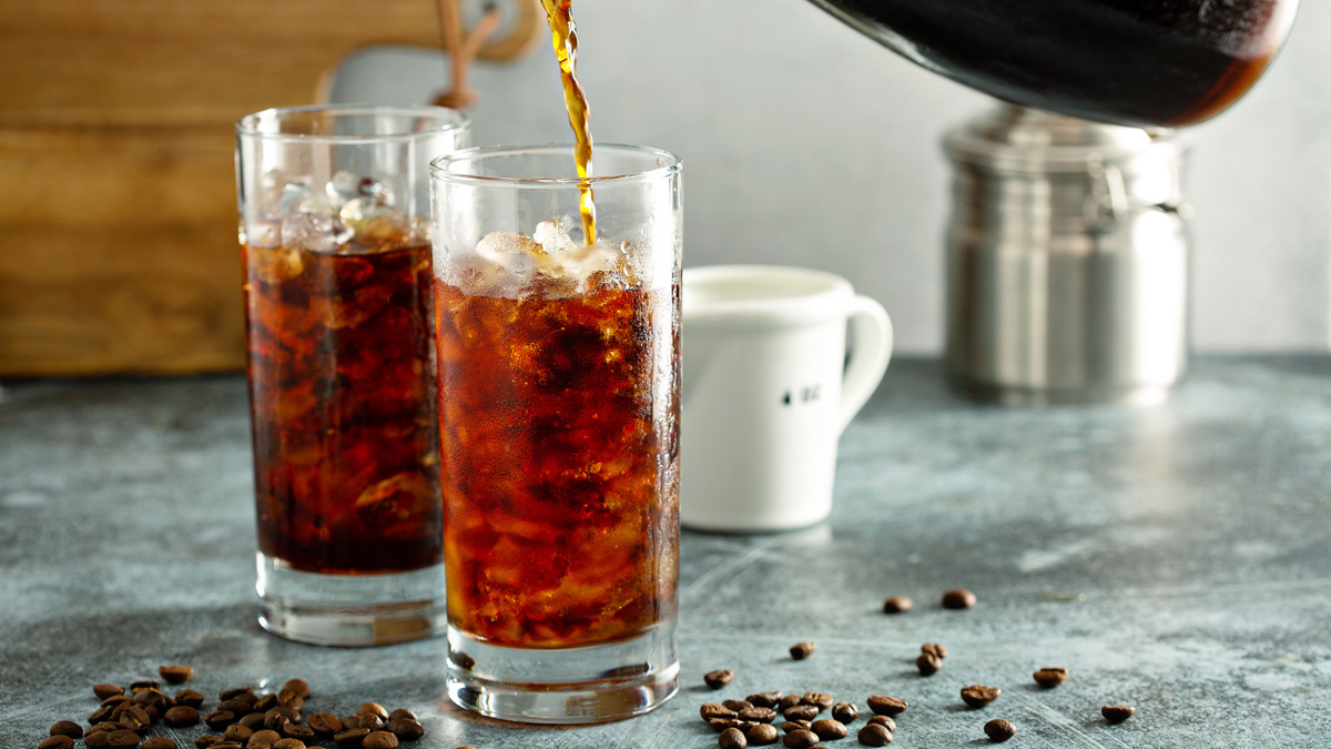 Cold brew being poured into a tall glass in front of an also full glass of cold brew. There are coffee beans scattered around on the counter.