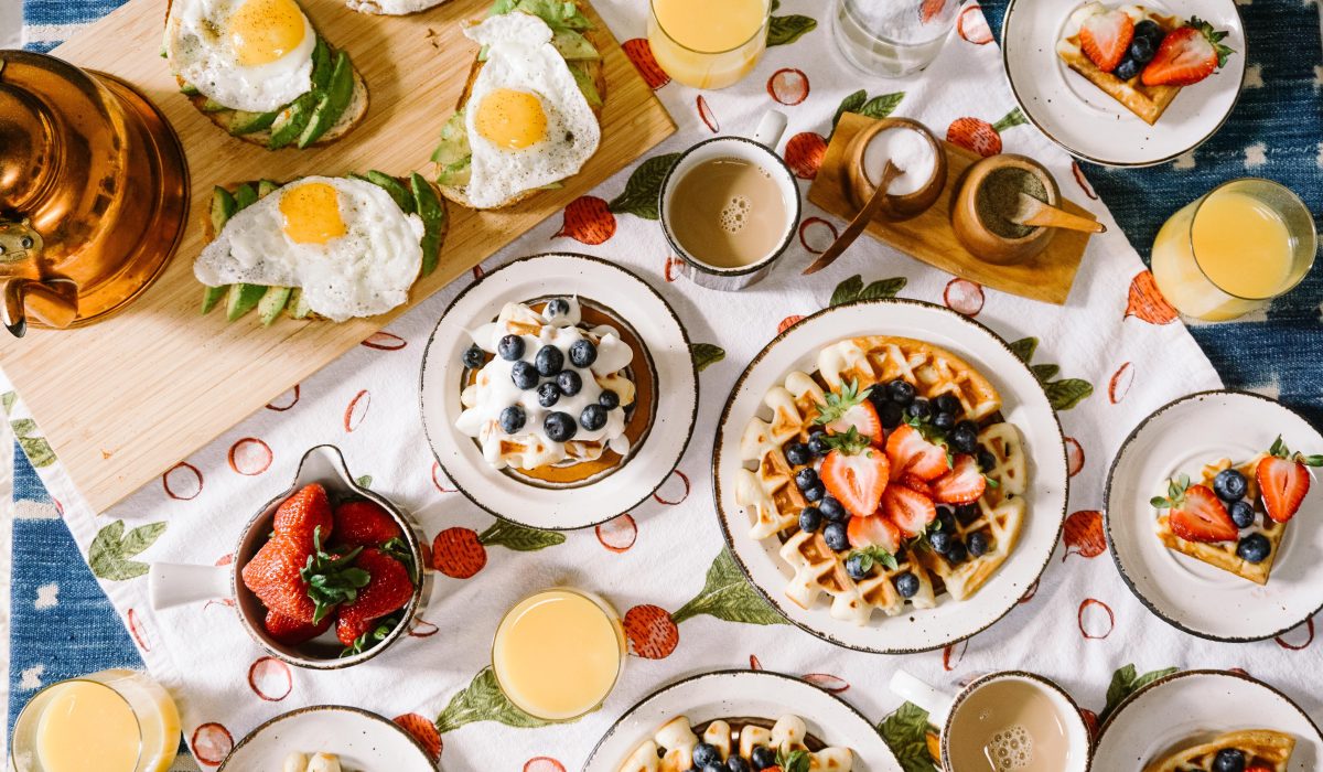 Kitchen table filled with breakfast foods - waffles, avocado toast with fried eggs, pancakes, fruits and coffee.