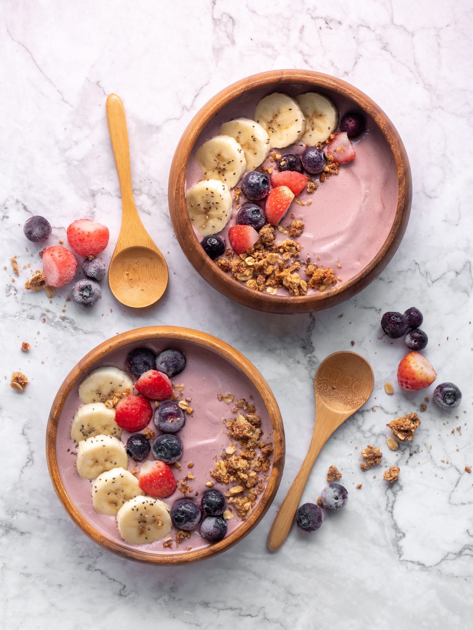 Pink-colored smoothies topped with bananas, berries and granola in 2 wooden bowls next to two wooden spoons.